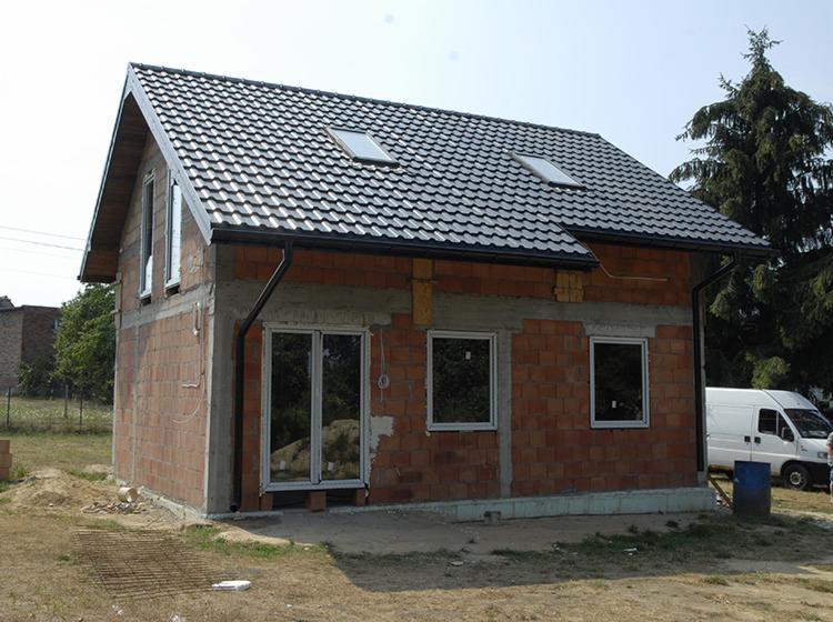 Niedokończony, dwukondygnacyjny budynek z czerwonej cegły z zamontowanymi oknami i pokrytym dachówką dachem z dwoma oknami dachowymi, widoczny z zewnątrz na tle nieba i drzew, z elementami...