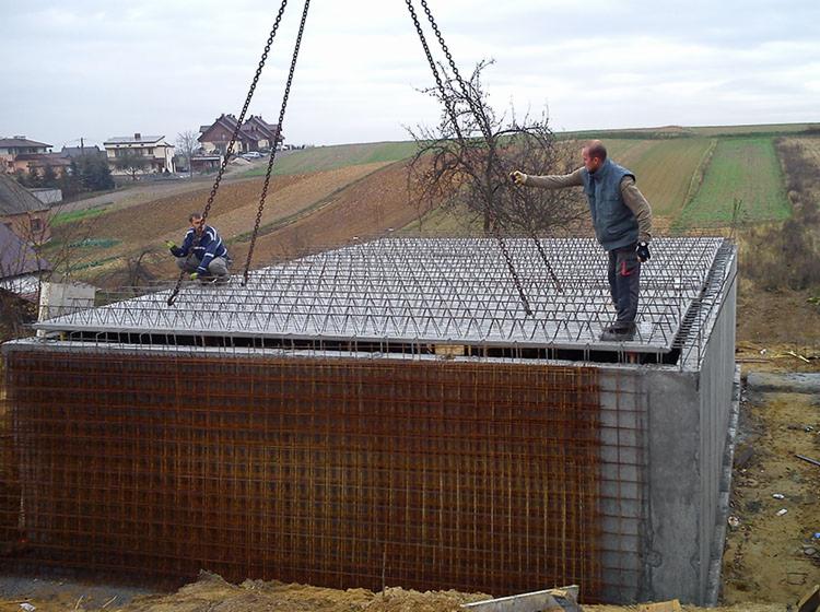 Budowa piwnicy: Dwóch pracowników montuje zbrojenie na betonowej konstrukcji, używając łańcuchów do podnoszenia elementów. W tle widoczne domy i pole uprawne.