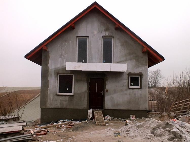 Surowy stan domu jednorodzinnego z czerwonym dachem, widoczne okna i drzwi wejściowe, elewacja w trakcie prac wykończeniowych, teren wokół w trakcie porządkowania, piwnica Kraków (według opisu).