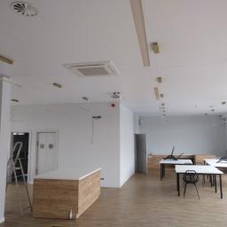 AKA KOMFORT - Wnętrze biurowe w trakcie wykończenia: widoczny sufit z centralną klimatyzacją kasetonową, okrągłe kratki wentylacyjne oraz wiszące lampy. Na ścianie widoczne niedokończone okablowanie. W tle...
