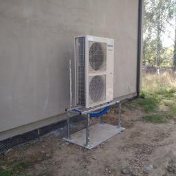 AKA KOMFORT - Zewnętrzna jednostka klimatyzacji Panasonic Inverter zamontowana na metalowej konstrukcji przy ścianie budynku z widoczną pianką montażową w szczelinie.