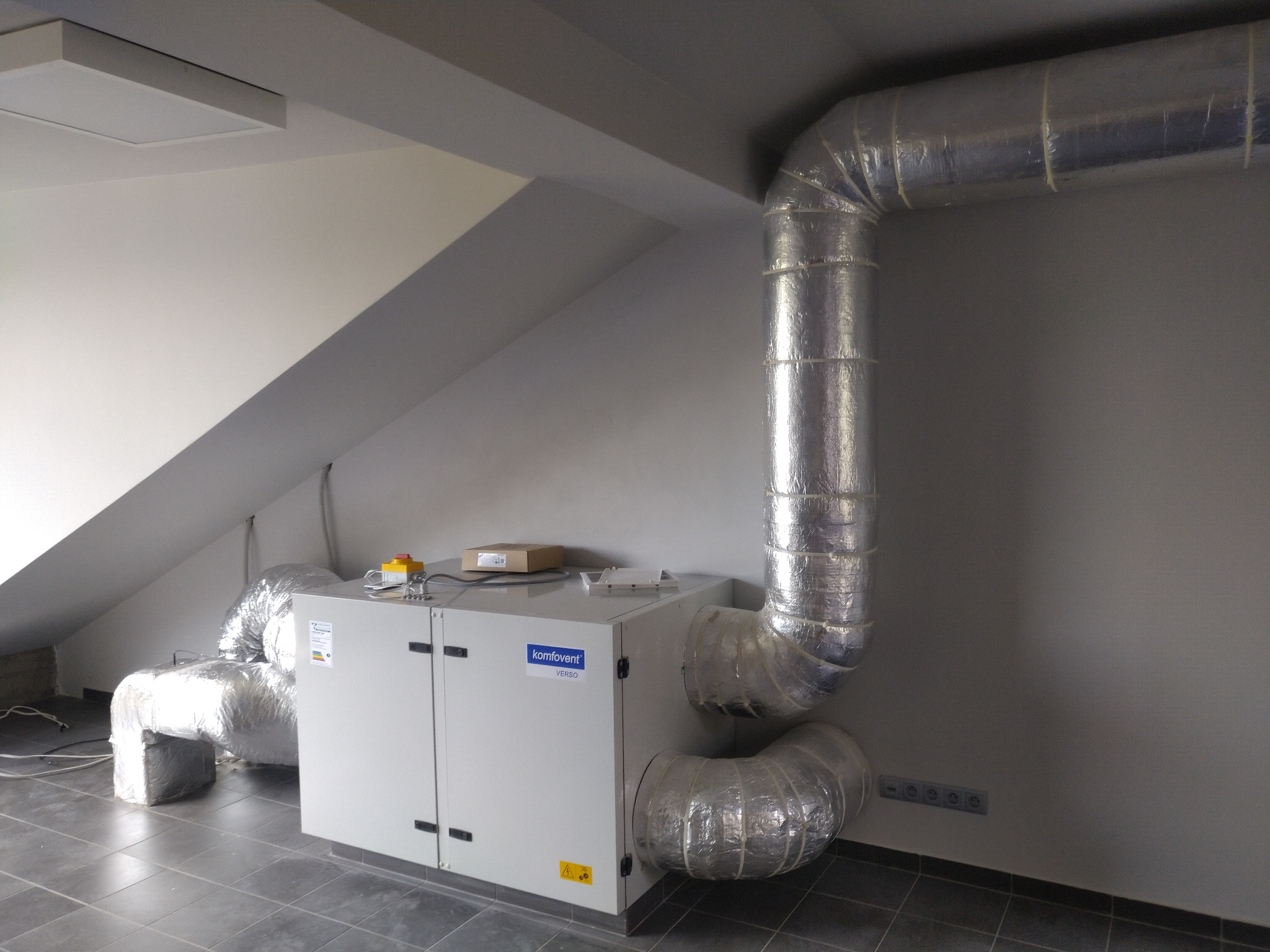 Szara instalacja wentylacyjna Komfovent Verso z izolowanymi rurami w pomieszczeniu na poddaszu w Raszynie, widoczne podłączenia elektryczne i skrzynka sterownicza.