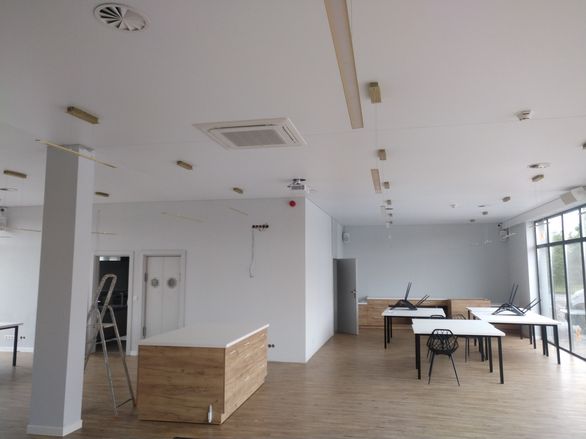 Wnętrze biurowe w trakcie wykończenia: widoczny sufit z centralną klimatyzacją kasetonową, okrągłe kratki wentylacyjne oraz wiszące lampy. Na ścianie widoczne niedokończone okablowanie. W tle...