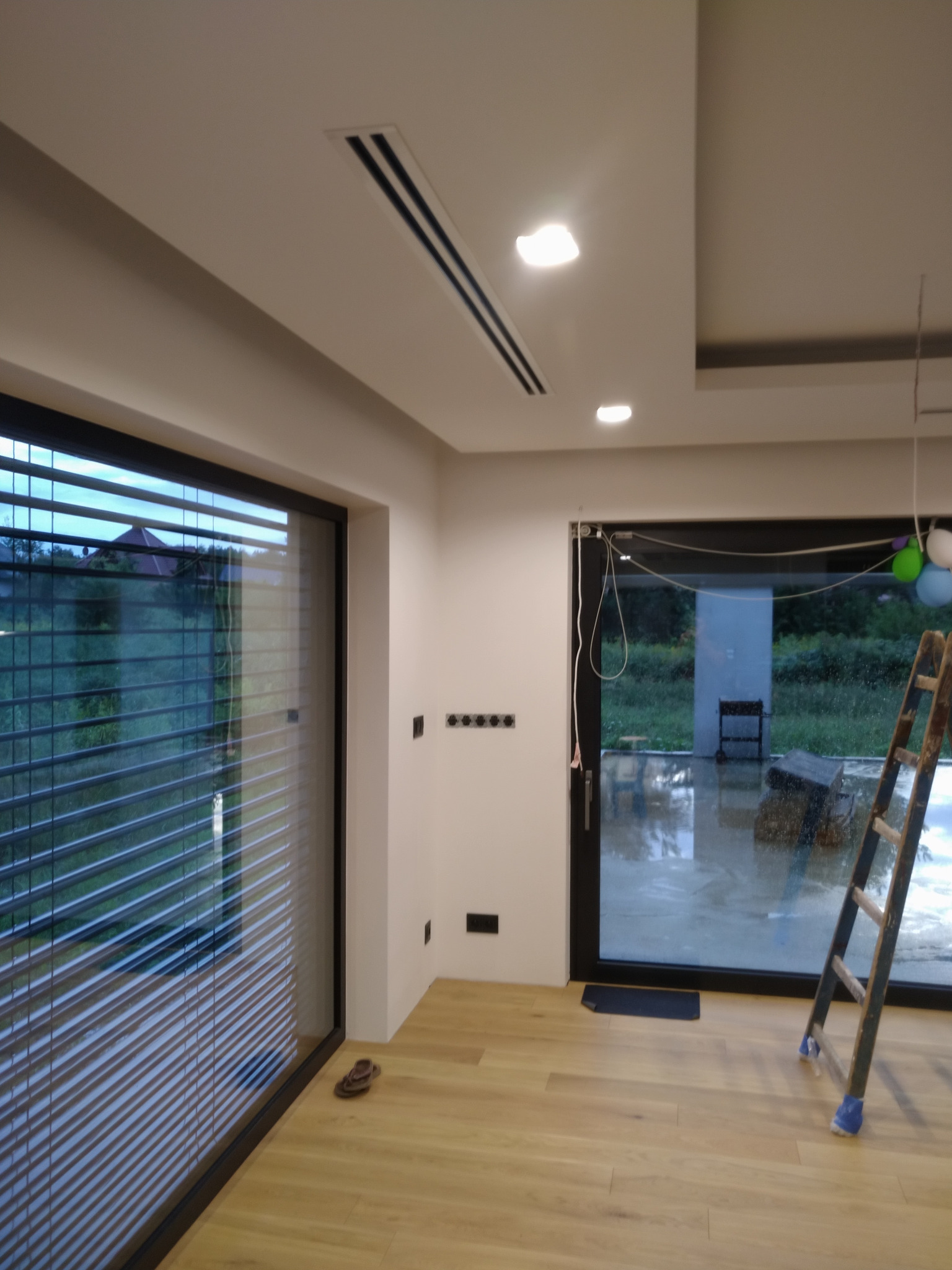 Wnętrze pokoju z zamontowanym anemostatem liniowym w suficie podwieszanym, widoczne duże okno z żaluzjami poziomymi, drabina oparta o ścianę i elementy instalacji elektrycznej.