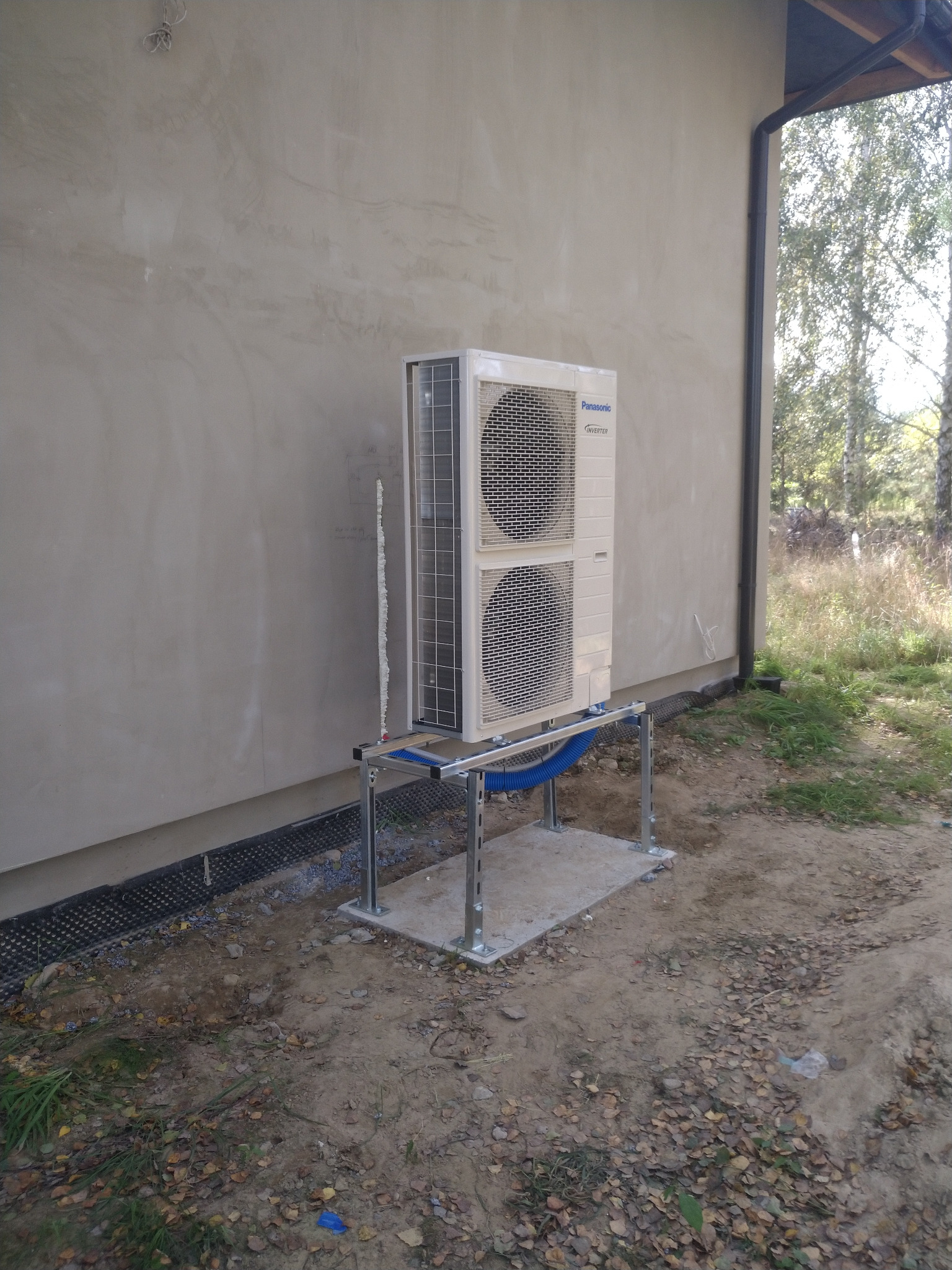 Zewnętrzna jednostka klimatyzacji Panasonic Inverter zamontowana na metalowej konstrukcji przy ścianie budynku z widoczną pianką montażową w szczelinie.