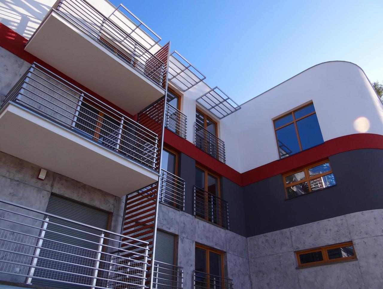 Nowoczesny budynek wielorodzinny z balkonami o metalowych balustradach, elewacja w kolorach szarości, bieli i czerwieni, okna z drewnianą ramą, widok z dołu na błękitne niebo.