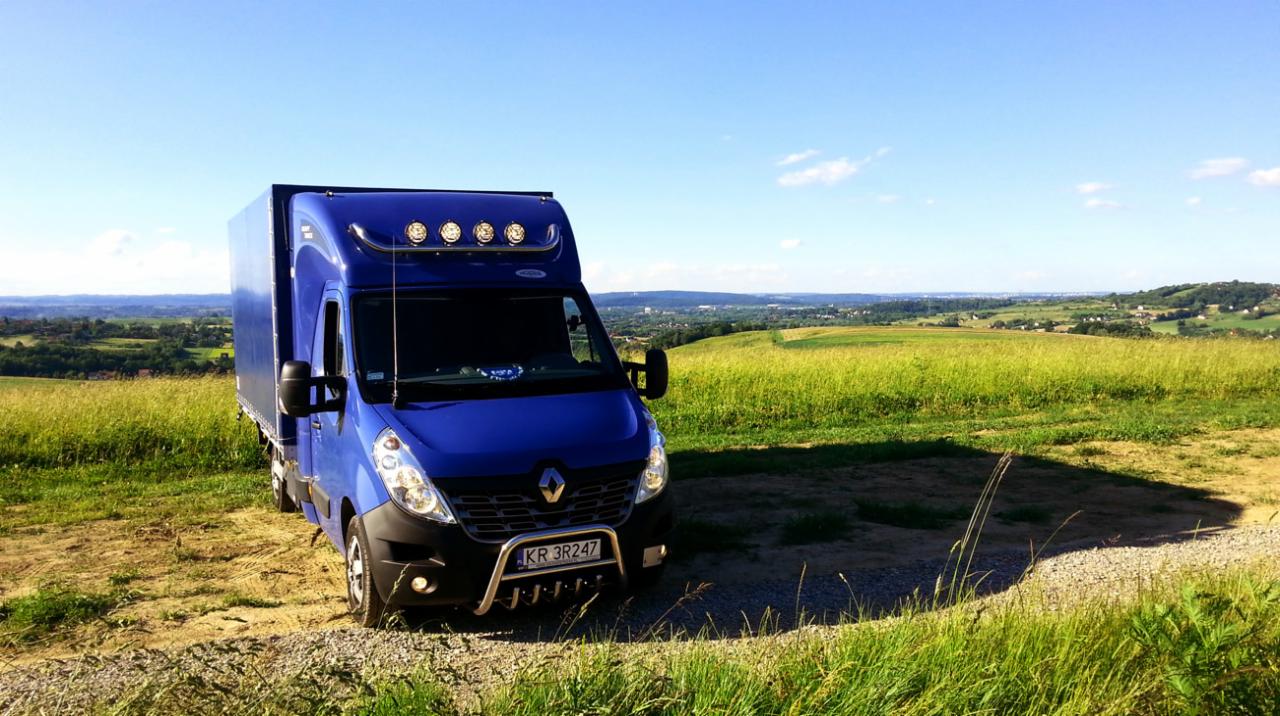 Niebieski samochód dostawczy Renault Master z zabudową kontenerową, zaparkowany na polnej drodze z widokiem na pagórkowaty krajobraz w tle.