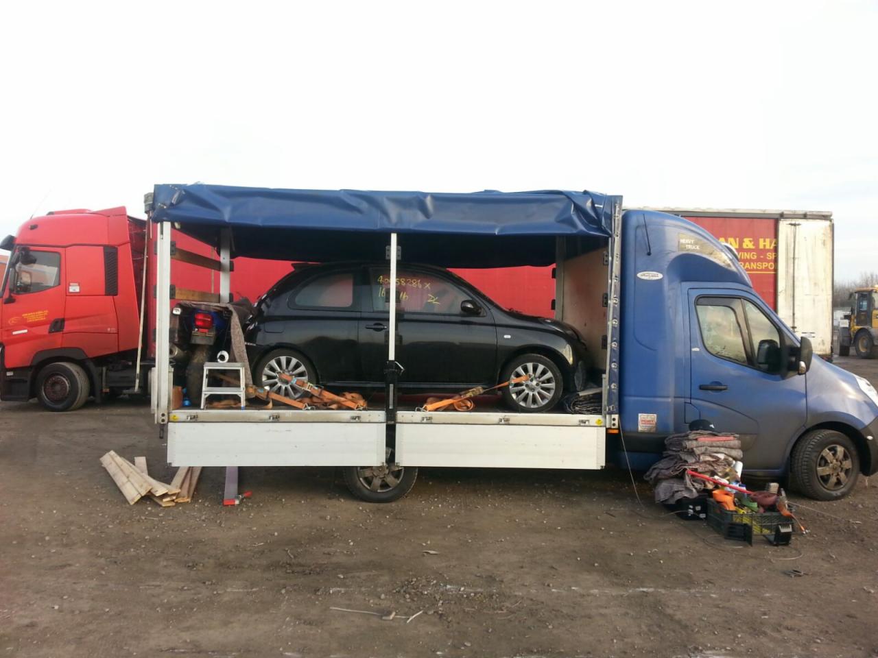 Czarny samochód osobowy zabezpieczony pasami transportowymi na platformie lawety z plandeką, obok czerwona ciężarówka i niebieski bus, teren piaszczysty.