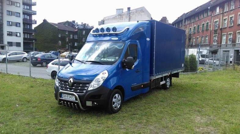 Niebieski samochód dostawczy Renault Master z plandeką, zaparkowany na trawie przed budynkami mieszkalnymi, z dodatkowymi lampami na dachu kabiny i orurowaniem z przodu.