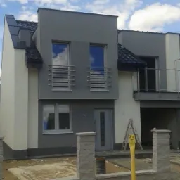 Dwukondygnacyjny dom jednorodzinny z elewacją w odcieniach szarości i bieli, widoczny fragment ogrodzenia z betonowych bloczków oraz żółty słup gazowy.