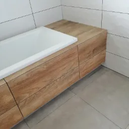 Nowoczesna łazienka z zabudowaną wanną w obudowie z drewnopodobnych paneli, białe płytki na ścianach z delikatnym wzorem, szare płytki podłogowe.