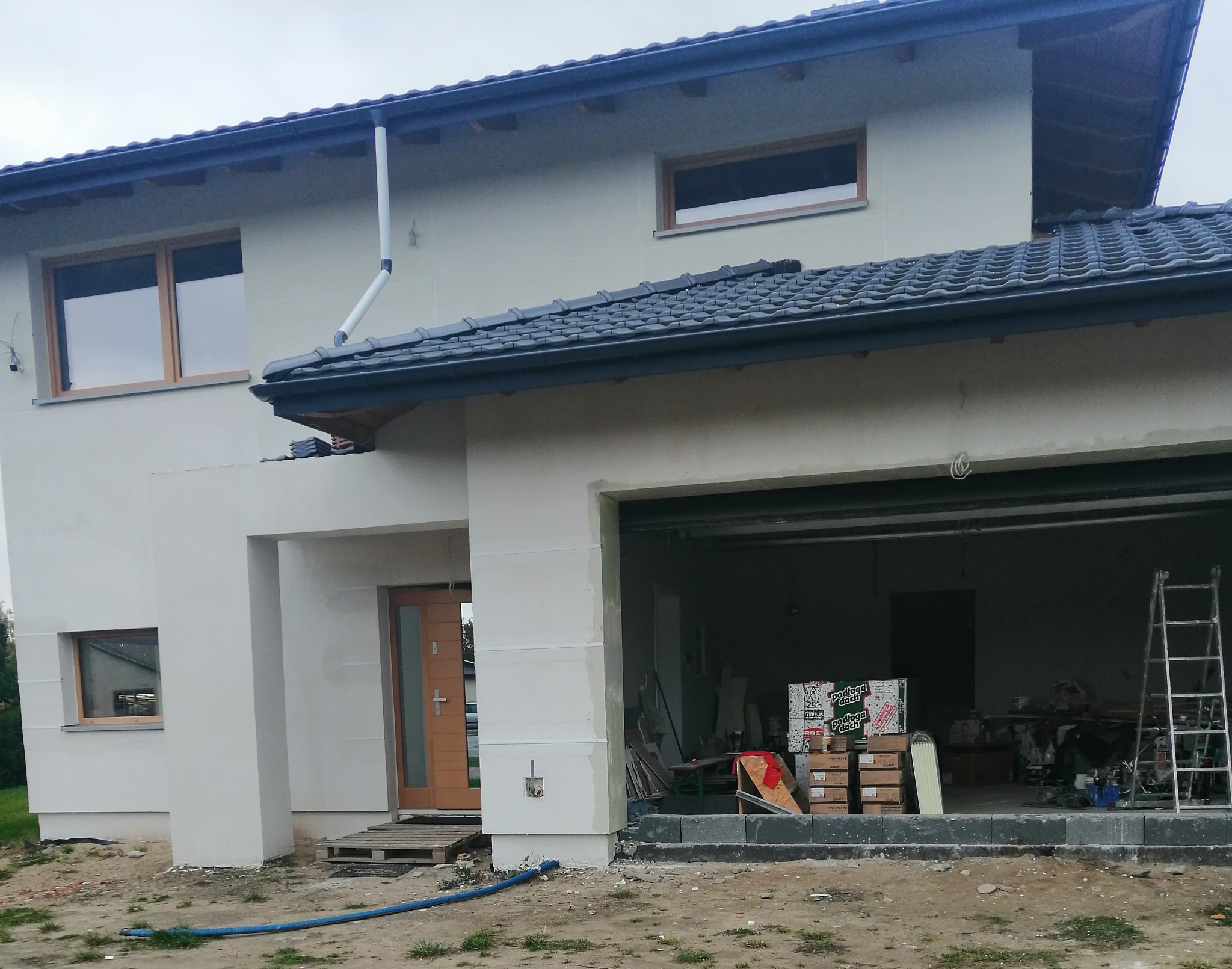 Dom jednorodzinny w trakcie tynkowania elewacji zewnętrznej, widoczne ślady szpachlowania i przygotowania pod ostateczną warstwę, z otwartym garażem i materiałami budowlanymi w środku.