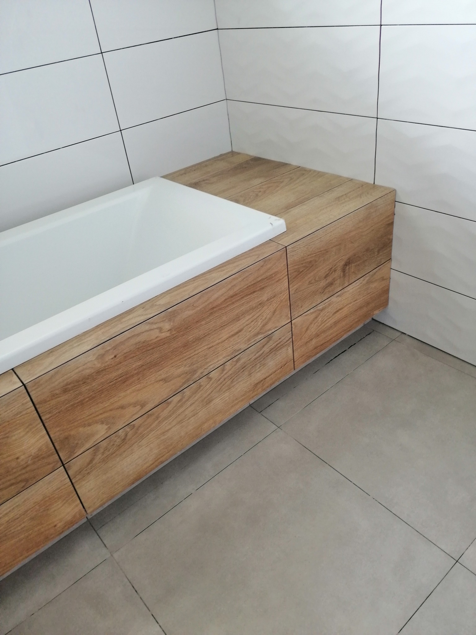 Nowoczesna łazienka z zabudowaną wanną w obudowie z drewnopodobnych paneli, białe płytki na ścianach z delikatnym wzorem, szare płytki podłogowe.