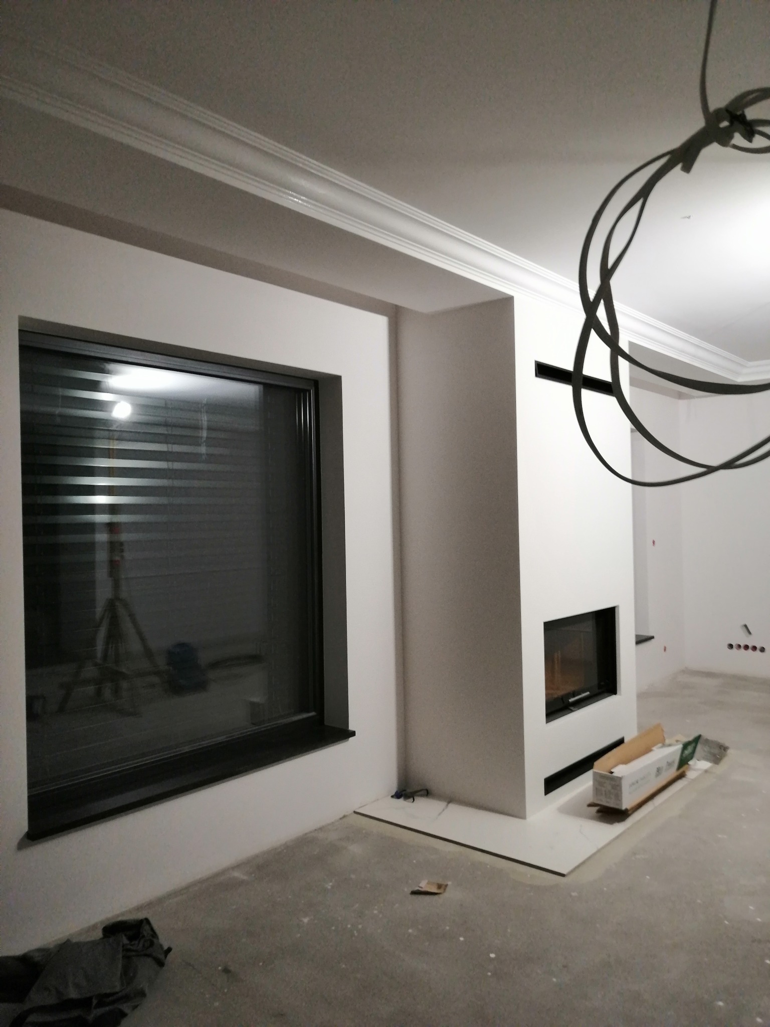 Wnętrze w trakcie wykańczania z nowoczesnym kominkiem, dużym oknem z żaluzjami i dekoracyjnymi listwami przysufitowymi. Na podłodze widoczne materiały budowlane.