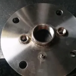 Okrągły, metalowy kołnierz z przyspawanymi gwintowanymi króćcami i otworami montażowymi leżący na metalowym blacie.