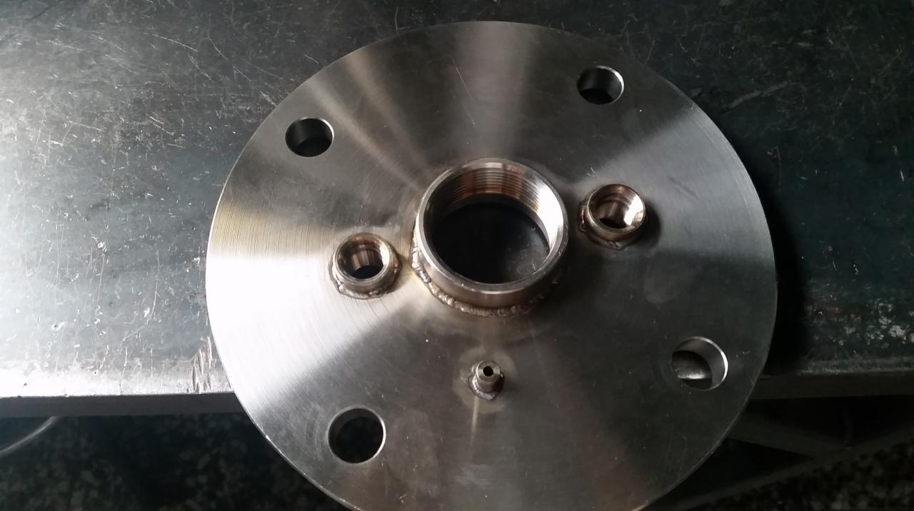 Okrągły, metalowy kołnierz z przyspawanymi gwintowanymi króćcami i otworami montażowymi leżący na metalowym blacie.