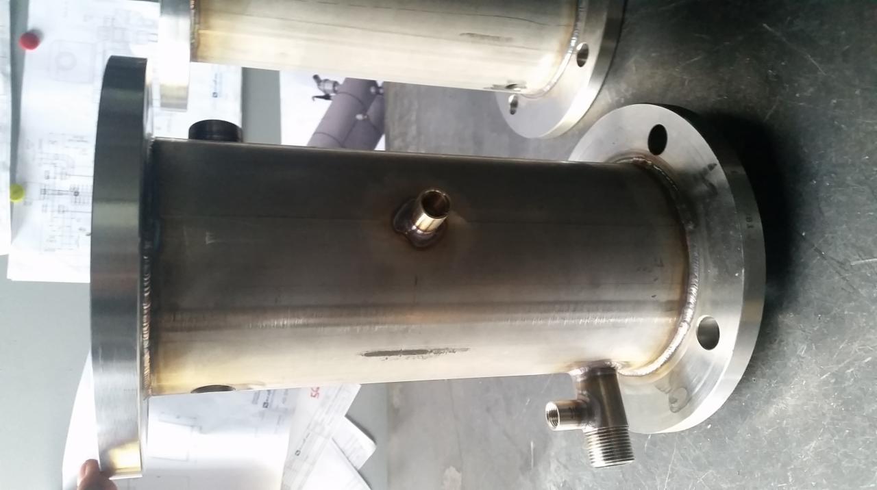 Dwa spawane, metalowe zbiorniki z kołnierzami, jeden na pierwszym planie z gwintowanym przyłączem, widoczne spawy i dokumentacja techniczna w tle.