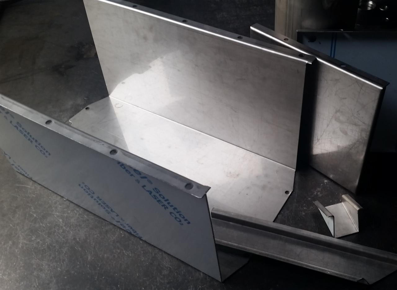 Metalowe elementy po obróbce, prawdopodobnie wycięte laserowo, z widocznymi otworami i zagięciami, leżące na ciemnym, metalowym stole warsztatowym.