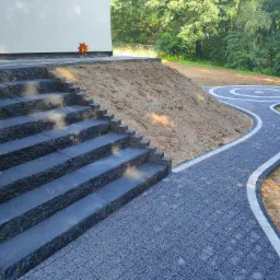 Schody z kostki brukowej prowadzące do domu, z ścieżką ułożoną z kostki brukowej w kształcie spirali, na tle zieleni.