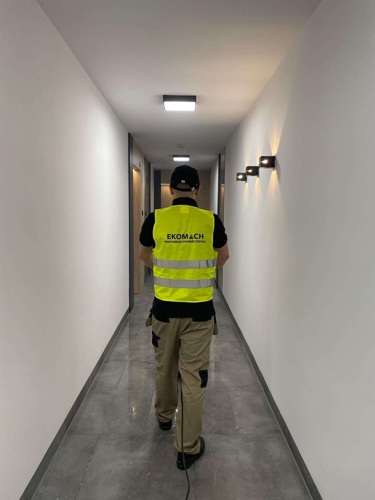 Pracownik w kamizelce firmy sprzątającej Ekomach myje podłogę w długim, jasnym korytarzu biurowym z nowoczesnym oświetleniem ściennym i podłogą z szarych płytek.