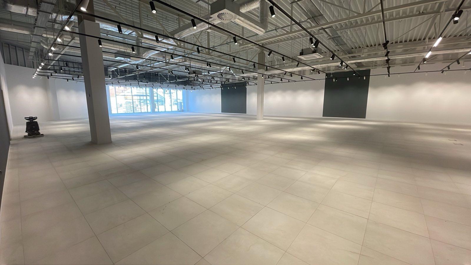 Wnętrze hali z jasną, kwadratową podłogą z płytek, oświetleniem szynowym i maszyną czyszczącą w rogu. Widok na okna i szare panele na ścianach.