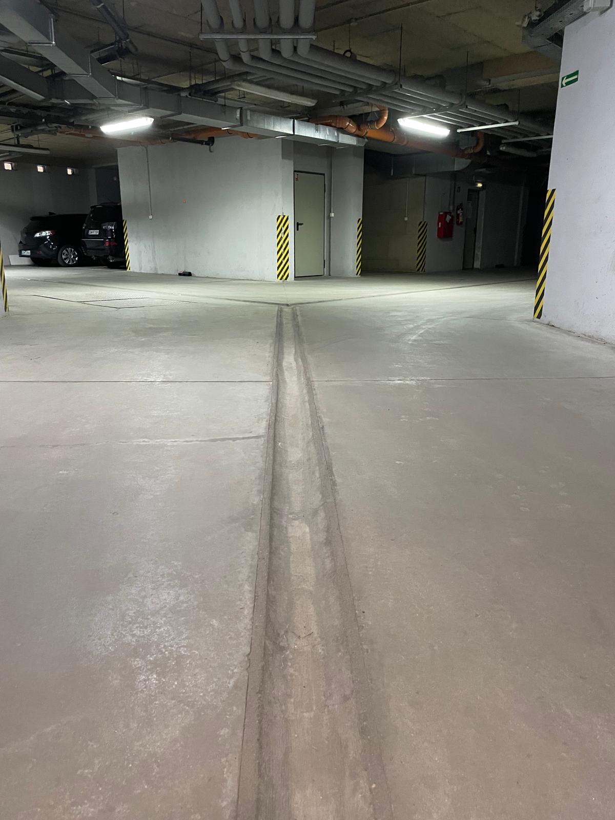 Czysta posadzka w garażu podziemnym z widocznymi liniami dylatacyjnymi i słupami oznaczonymi pasami ostrzegawczymi. W tle zaparkowane samochody.