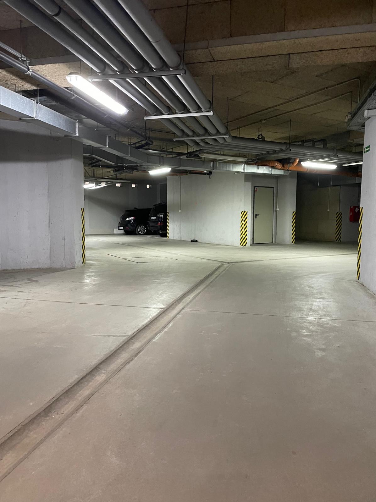 Czysty, betonowy parking podziemny z widocznymi instalacjami pod sufitem, oświetleniem i dwoma zaparkowanymi samochodami w tle. Słupy oznaczone pasami ostrzegawczymi.