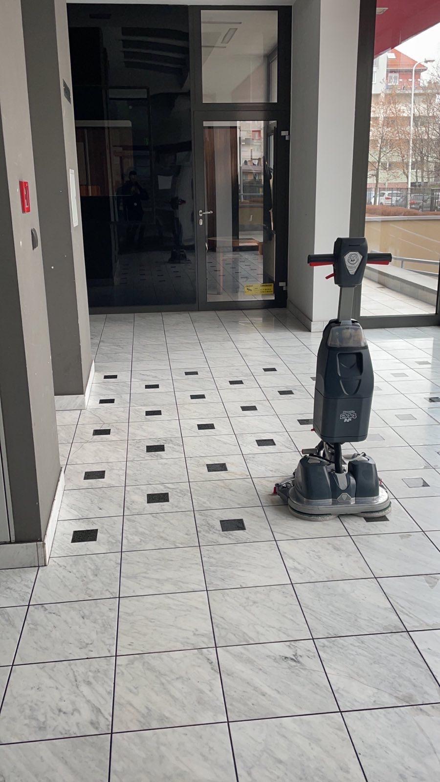 Automat szorująco-zbierający do mycia podłóg na marmurowej posadzce w holu biurowca. Widoczne drzwi wejściowe i fragment budynku w tle.