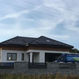 Dom jednorodzinny z panelami słonecznymi na dachu, widoczny firmowy bus z napisem 'Instalacje Fotowoltaiczne', ogrodzenie z szarej kostki.