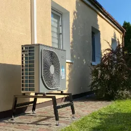 Zewnętrzna jednostka pompy ciepła zamontowana na metalowej konstrukcji z regulowanymi nóżkami, ustawiona na kostce brukowej przy ścianie budynku o żółtej elewacji, w tle widoczne okna i zieleń ogrodu.