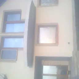 Fragment elewacji budynku z oknami o różnej wielkości, z elementami dekoracyjnymi w postaci listew wokół okien i łukowego daszku nad jednym z nich, ściana w jasnym odcieniu beżu.