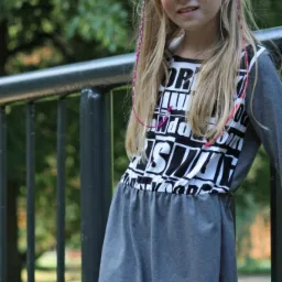 Dziewczynka w szarej sukience z czarno-białym nadrukiem, oparta o metalową barierkę na tle zieleni.