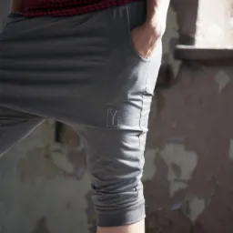 Szare spodenki dresowe 3/4 z logo marki na udzie, kieszeń z przodu, osoba w czerwono-czarnej koszuli w kratę, na tle ściany z łuszczącą się farbą.