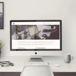 Monitor iMac wyświetlający stronę internetową z minimalistycznym designem, obok filiżanka, roślina w doniczce i zdjęcie górskie w ramce, na białym biurku.