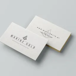Dwie wizyt&oacute;wki firmy Marine Gold z Kalwarii Zebrzydowskiej, z minimalistycznym logo przedstawiającym żagl&oacute;wkę, na jasnoszarym tle. Jedna wizyt&oacute;wka leży płasko, a druga jest ustawiona pod kątem...