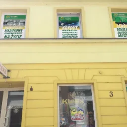 Żółty budynek z trzema oknami na górze, w których wiszą plakaty reklamowe z informacjami o ubezpieczeniach, kredytach i leasingu, oraz szyld 'KLEKS' nad wejściem.