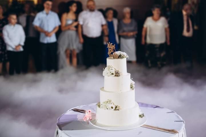 Czteropiętrowy tort weselny z białym kremem i żywymi kwiatami, zwieńczony napisem 'Miłość jest słodka', na stole otoczonym sztuczną mgłą, w tle goście.