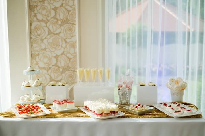 Elegancki stół z deserami: mini bezy, kremowe desery w szklankach, ciastka na paterze i w kieliszkach, dekoracje kwiatowe, złoty bieżnik, jasne tło z firanką i wzorem w róże.
