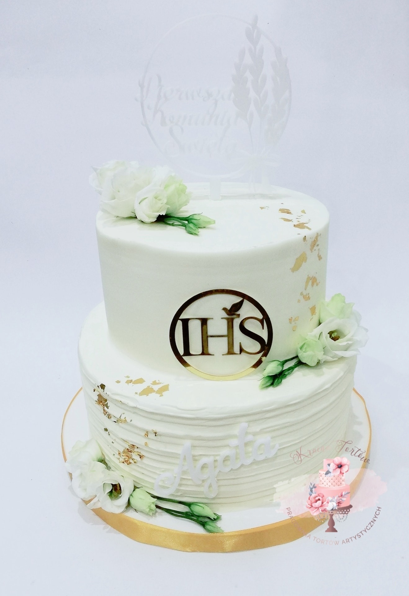 Dwupiętrowy tort komunijny w białej polewie, zdobiony złotymi płatkami, białymi kwiatami eustomy, złotym napisem IHS z gołębiem i topperem z napisem 'Pierwsza Komunia Święta' oraz imieniem 'Agata'.
