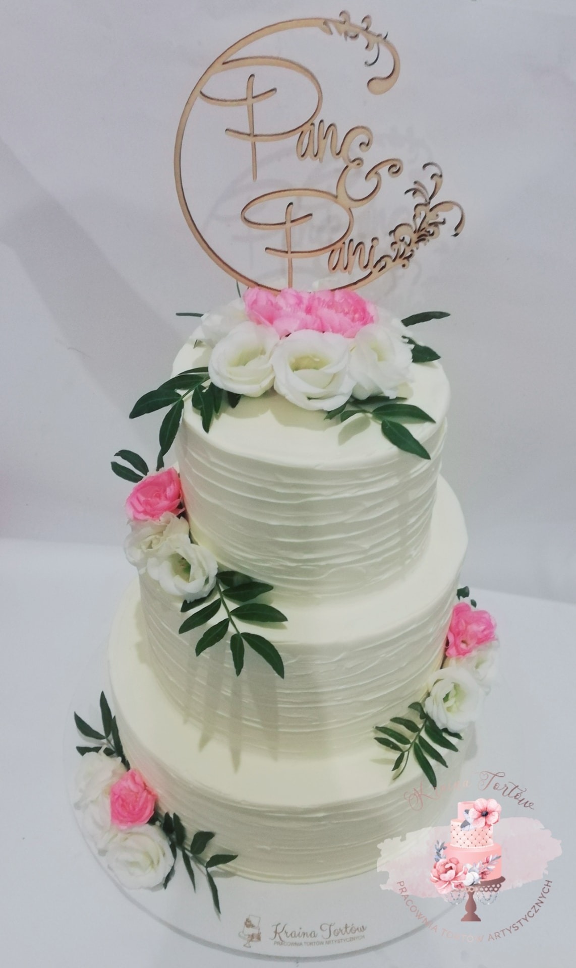 Trzypiętrowy tort weselny z kremowym lukrem, zdobiony białymi i różowymi kwiatami oraz drewnianym topperem z napisem 'Państwo Państwo'.