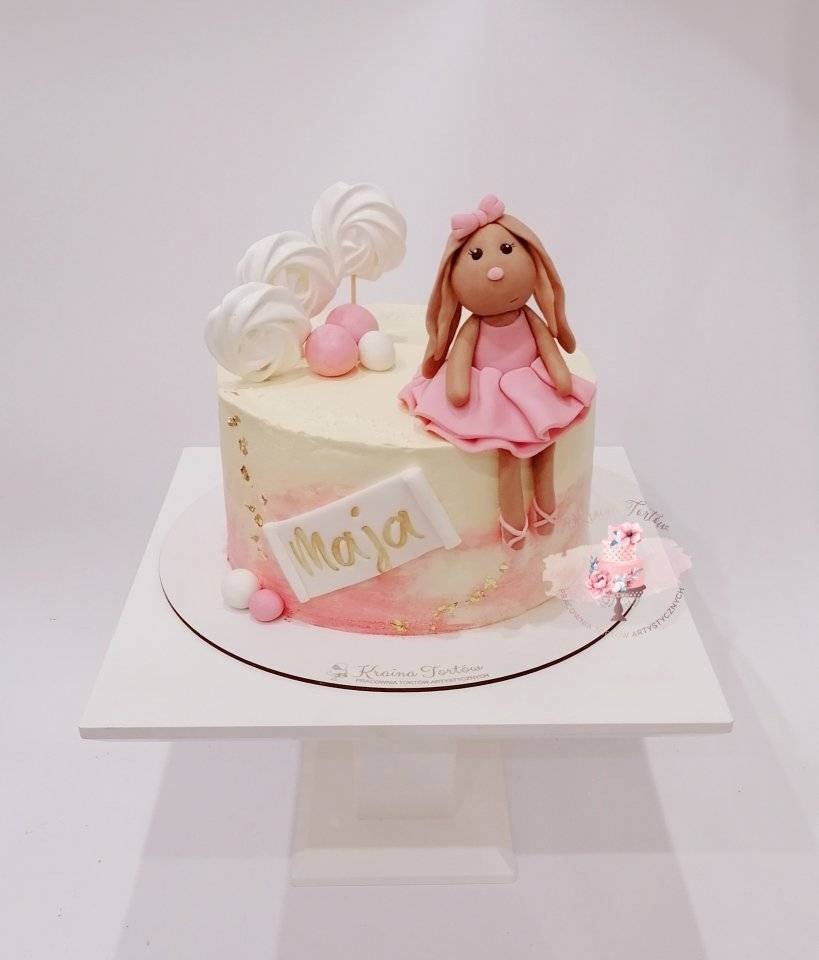 Tort z kremowym wykończeniem, różowymi akcentami i figurką lalki w różowej sukience z kokardą na głowie; dekorowany bezami na patyku i napisem 'Maja' wykonanym złotą czcionką.