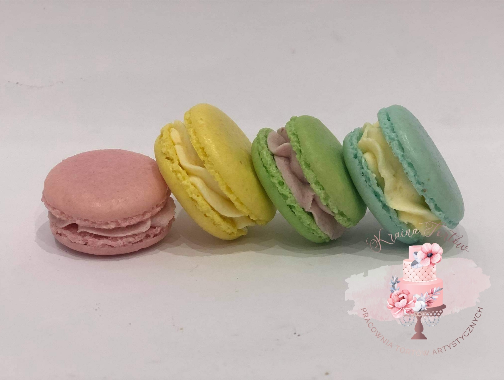 Rząd kolorowych makaroników: różowy, żółty, zielony i turkusowy, ustawionych pod kątem na białym tle, z logo cukierni w prawym dolnym rogu.
