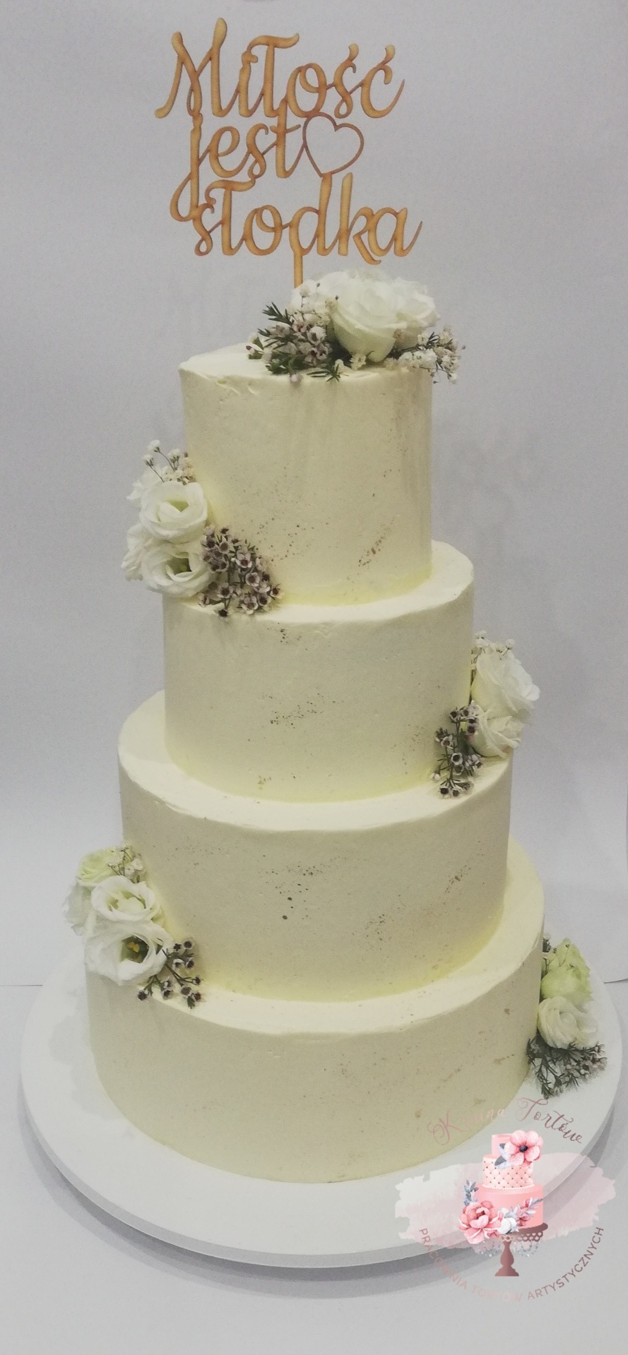 Elegancki, czteropiętrowy tort weselny pokryty kremowym lukrem, ozdobiony białymi kwiatami i drewnianym topperem z napisem 'Miłość jest słodka'.