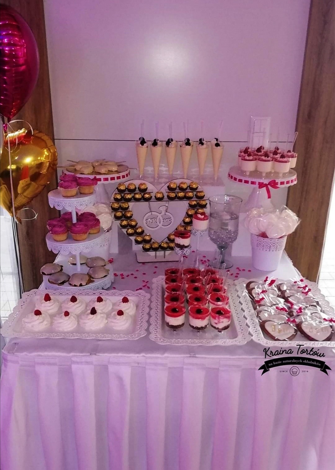 Stół z deserami na przyjęciu: babeczki, mini desery w pucharkach, rożki z kremem, praliny w kształcie serca z napisem 'Mr & Mrs', ciasteczka w kształcie serca z lukrem, balony w kolorze różowego...