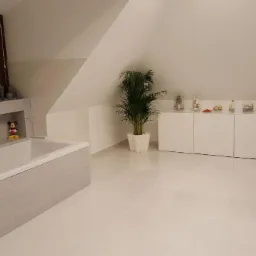 Jasna łazienka na poddaszu z białą wanną obudowaną szarymi płytkami, białą szafką, umywalką z ciemną szafką i wysoką rośliną doniczkową; na wannie dekoracje i figurka Myszki Miki.