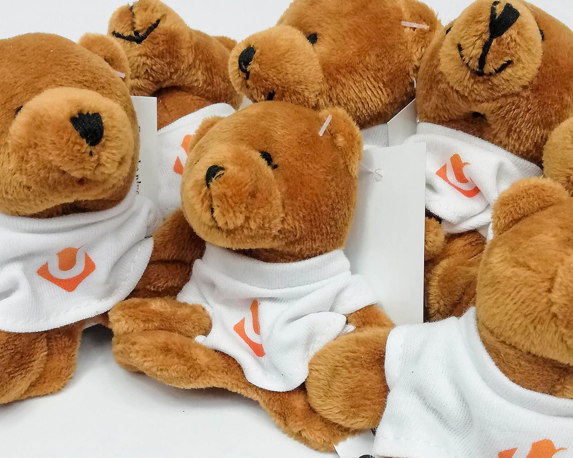 Pluszowe misie w białych koszulkach z pomarańczowym logo, ustawione blisko siebie na białym tle.