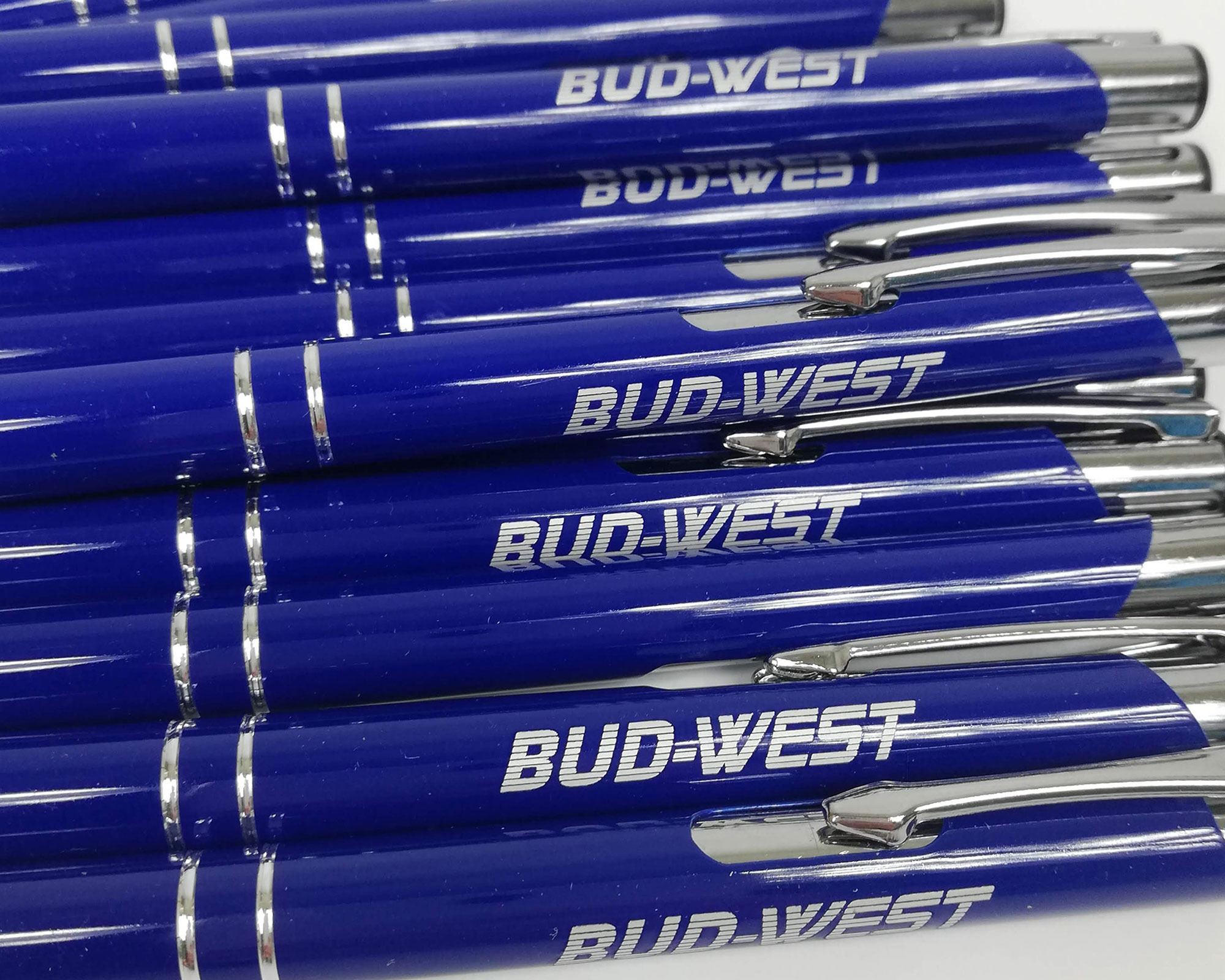 Stos niebieskich długopisów z nadrukowanym białym logo firmy BUD-WEST, ułożonych równo, z widocznymi srebrnymi elementami.