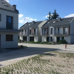 Nowoczesne, dwukondygnacyjne domy w zabudowie szeregowej z balkonami i szarą dachówką, widziane w słoneczny dzień, z częściowo zagospodarowanym terenem wokół.