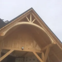 Wnętrze drewnianej konstrukcji domu w trakcie budowy, widoczny łukowy element nośny, surowe drewno, ściany w stanie deweloperskim, drabina i lampa na suficie.