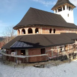 Budowa kościoła w zimowej scenerii, widoczne rusztowania, ceglana konstrukcja i czarny dach z wieżą w tle.